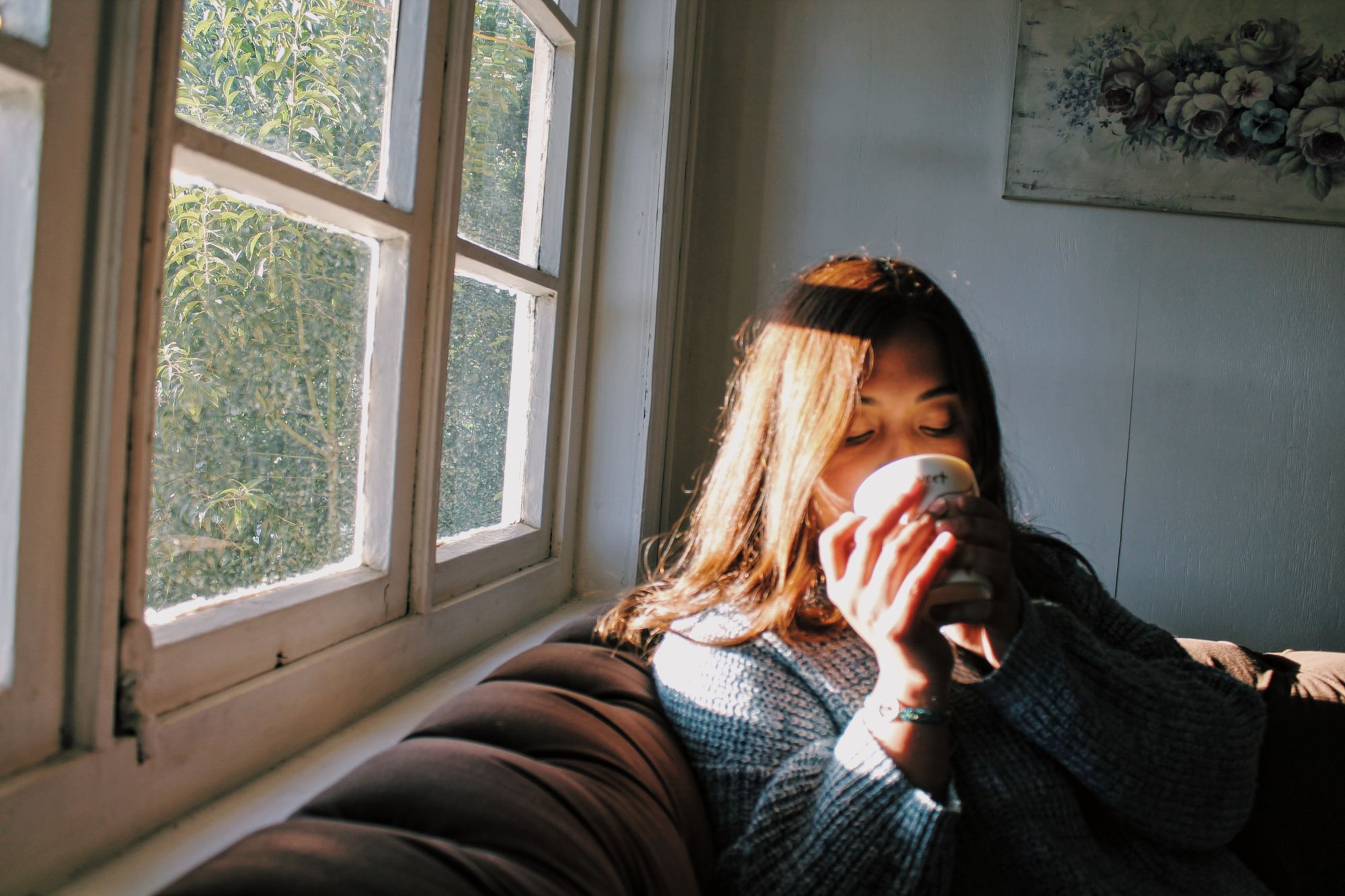 Mulher tomando uma bebida quente no sofá, junto à janela, com o sol no rosto.