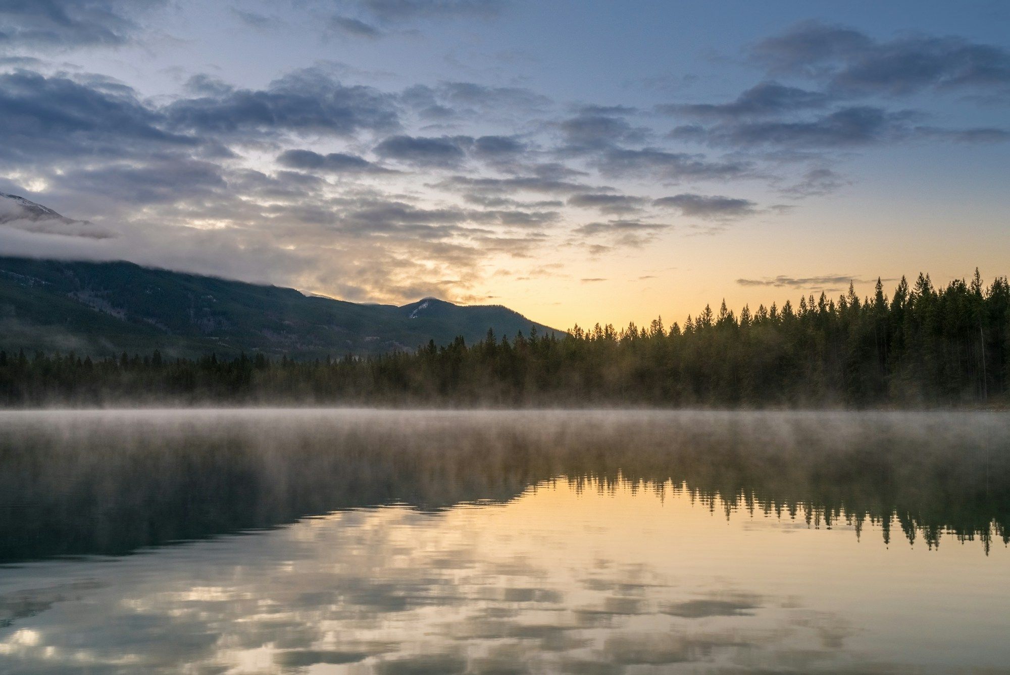 Lago parado com névoa ao nascer do sol, com montanhas e floresta refletidas.